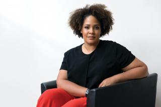 Ijeoma Oluo sits in a black leather chair, wearing a black shirt and red trousers.