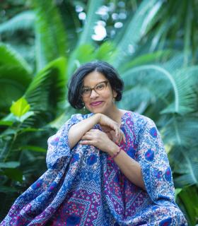 Rev. Mariela, in a brightly colored caftan, sits smiling in front of a lush wall of green foliage.