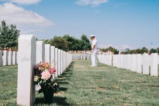 A man in US Navy dress whites stands on the grass in a military cemetery looking at the headstones