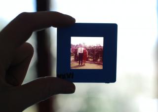 A hand holds a slide up to the light, on which is a dated image of a woman and man posing outside.