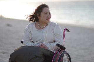 An Asian woman sits in a pink wheelchair outdoors, perhaps in front of a body of water. She is wearing an ivory sweater and a pearl necklace.
