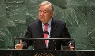 UN Secretary-General António Guterres is seen speaking at the podium of the UN General Assembly Hall. He is wearing a black suit with a red tie.