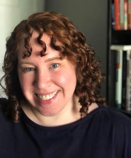 white woman with reddish brown curly hair, wearing a dark blouse, in front of a bookcase
