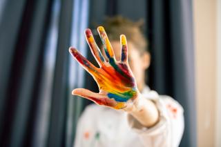 A person holds their palm to the camera, obscuring their face. Their palm is painted with streaks of bright paint in yellows, reds, and blues.
