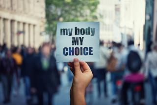 A white person's hand holding a sign with the message "my body my choice" over a crowded street