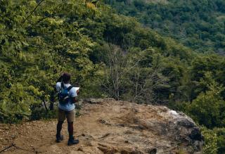 On a rocky ledge overlooking the wildnerness, a Black man wearing a backpack holds a map.