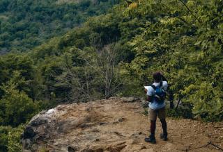 On a rocky ledge overlooking the wilderness, a Black man wearing a backpack holds a map.