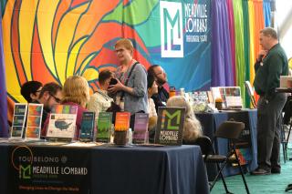 A group of people at a booth in the GA exhibit hall. Pamphlets are displayed on the table, and a the exhibitor displays a colorful backdrop company logo behind their table. Other colorful drapes are hung in the adjacent booth.