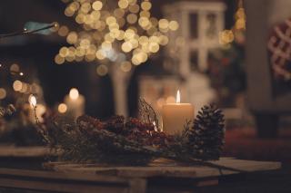 A white candle burns, in a dim room, resting near evergreen and pinecone. Twinkly lights are visible in the background.