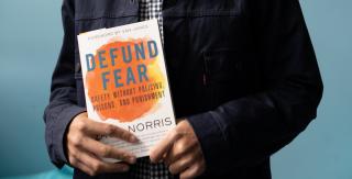 A book titled "Defund Fear" by Zach Norris is held in the hands of someone whose hands are light brown; only their torso is showing.
