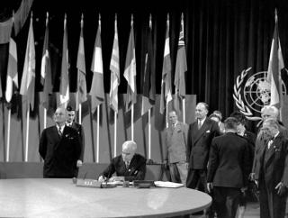 Black and white photo of the signing of the United Nations Charter in a room lined with national flags from UN member states and the UN logo on a curtain to the right. US Secretary of State Edward R. Stettinius, Jr. sits at a round table to sign the document while President Harry S. Truman stands by at the left looking pleased. Additional diplomats stand behind wearing suits.