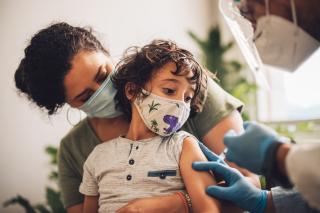 A child, wearing a mask and sitting on their parent's lap, is given a vaccine by a health care provider in mask and gloves.
