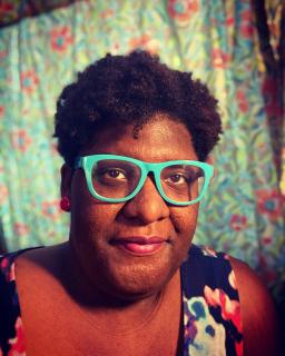 JeKaren, a Black woman with short hair, wears red earrings, pink lipstick, and glasses with bright teal frames.