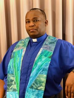 Rev. Ali stands, wearing a blue clergy shirt and quilted stole, with a somber and fierce expression on his face.
