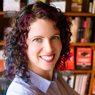 Sara Palmer smiles broadly, in front of a bookcase. She has curly, brown hair with pink and blue streaks.