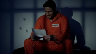 A man in an orange prison jumpsuit reads a letter, smiling.