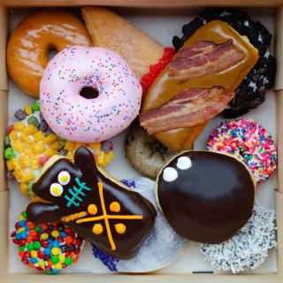 A vibrant box of colorful donuts from Voodoo Donuts in Portland, OR. One donut includes pieces of bacon and maple frosting. Another donut is painted to look like a "Voodoo doll". 