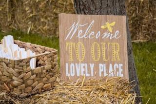 A wooden sign stating "Welcome to Our Lovely Place" leaning against a tree with a picnic bask