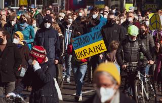 Crowd of peaceful protestors wearing masks; a prominent sign reads "Stand With Ukraine."