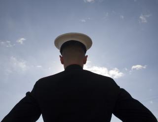 From behind, the silhouette of a U.S. Marine in dress uniform with a blue sky and a few clouds