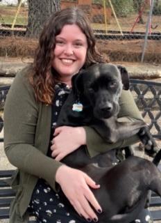 Jaimie Dingus, wearing a green sweater and black dress, holds her black dog in her lap as she sits on a park bench