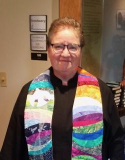 a person with short down hair wears a black robe and colorful stoll.