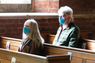 Two women, each wearing a mask, sit in wooden church pews.