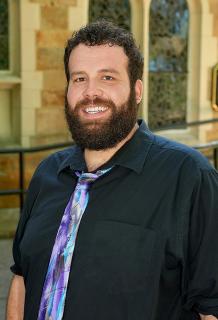 man with beard, in a dark shirt and purple/blue tie, smiles in front of a church