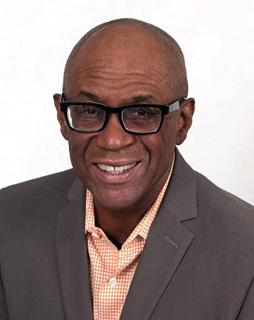 black man wearing a gray suite, peach shirt, and black glasses, smiles