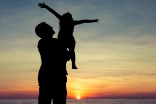 Against a sunset, an adult holds up a child with their arms outflung joyfully
