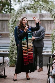 Erika wears a black clergy robe and a rainbow stole. She is speaking into a microphone and standing in front of two men mid-wedding ceremony.
