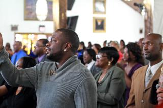 Inside a church, Black people stand as if listening to a preacher or choir.