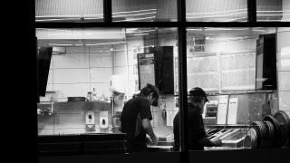 A candid black-and-white shot at night, looking through window of nearby pizza parlour. Two workers are seen from behind as they prepare food.