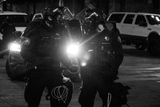 Grayscale photo of two police officers in black helmets, black gas masks, and black riot gear, with zip ties on their belts and nightsticks in their hands, standing in front of several SUVs