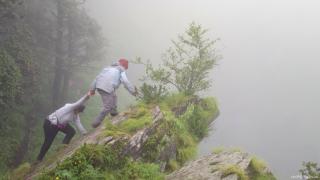 On a rocky outcrop in what appears to be inclement weather, one person strenuously pulls another person uphill.