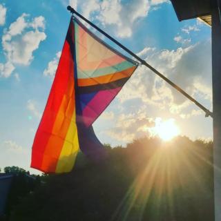 A Progress Pride flag on a flagpole, with a blue sky and bright sun behind it.