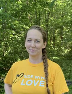 image of a person with hair in a braid wearing a yellow Side With Love shirt, standing in front of green trees