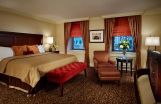A photo of a guest room at the Omni William Penn hotel. The room features a single king sized beds with tan bedding and a padded red bench. There is also an upholstered chair sitting infront of two windows with long drapes. istance.