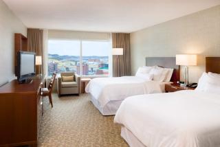 A photo of a guest room at the Westin hotel. The room features a flat screen TV and two queen sized beds with white bedding. A view of the yellow Fort Pitt bridge can be seen in the distance.