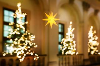 A lit Moravian star lamp hangs in what appears to be a church, with a few lit Christmas trees blurred in the background.
