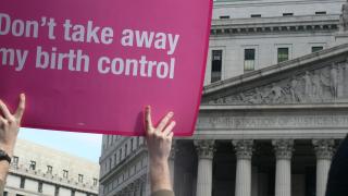 A pink placard reading "Don't take away my birth control" taken at a Planned Parenthood Rally in New York City. In the background, the stonework of a courthouse is visible.