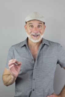 Eric Lane Barnes, a white man with trim white beard in posed to conduct, wearing a flat "ascot" cap and gray collared shirt