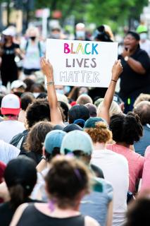 A crowd of protestors, from behind. One person holds aloft a sign, "Black Lives Matter," while in the background a Black person uses a microphone