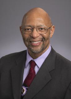 A bald, brown skinned man with a beard and glasses, wearing a suit jacket with dark red tie.