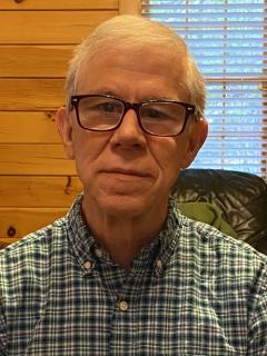 A white man with silver hair and glasses wears a green plaid shirt.