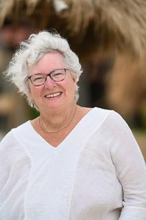 Sue, a white woman with short white hair, smiles outdoors. She's wearing a white top and black-rimmed glasses.