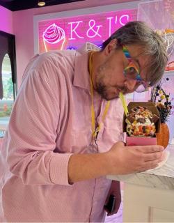 A person with light brown and gray hair and beard, wearing rainbow colored glasses and a pink shirt poses with a cupcake in front of a neon pink sign of a cupcake.