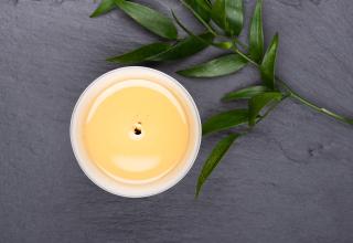 Seen from above, an ivory pillar candle burns on a slate surface with a branch of greenery resting nearby.