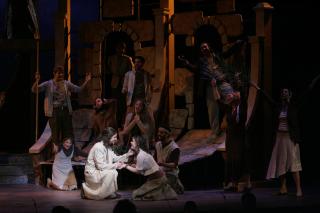 The cast of "Jesus Christ Superstar" on stage, at a moment where Jesus is kneeling with a woman and smiling.