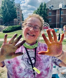 smiling person with glasses, a paint-covered tie dye shirt and paint covered hands with greenery behind